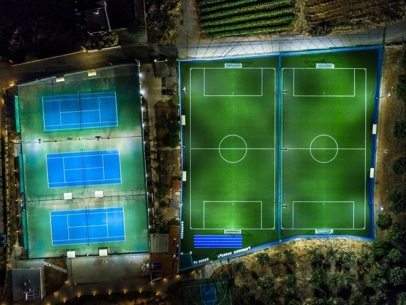 Vista nocturna de un complejo deportivo con pistas de tenis iluminadas y campos de fútbol.
