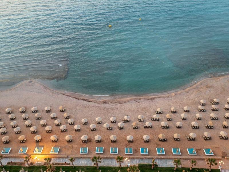 Vista aérea de una playa con sombrillas y tumbonas junto a un mar tranquilo y claro.