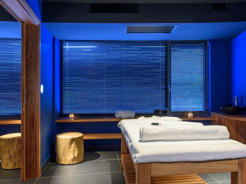 Sala de masajes acogedora con banco de madera, taburetes y camilla con sábanas blancas bajo luz tenue.