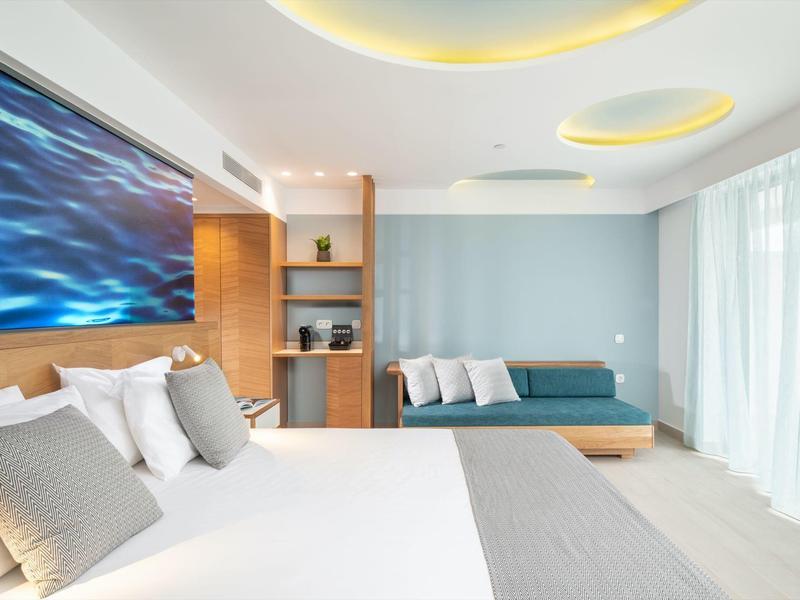 Habitación de hotel moderna con cama grande, área de sofá, estantes de madera y ventana grande con cortinas.