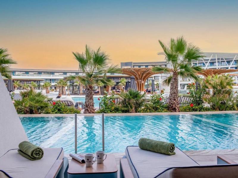 Piscina del hotel con tumbonas y toallas verdes, rodeada de palmeras y vegetación bajo un cielo al atardecer.