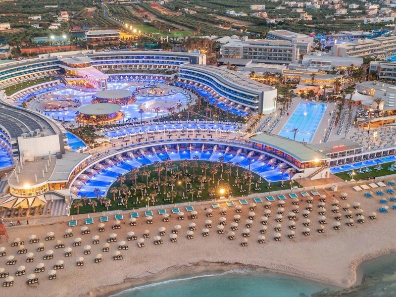 Gran complejo hotelero con piscinas y playa iluminado de noche.