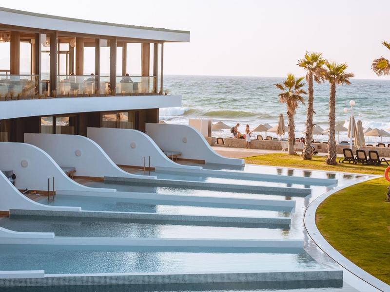 Piscina moderna de hotel con tumbonas integradas y vista al mar y palmeras.