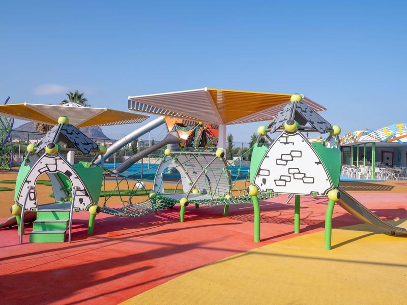 Juegos infantiles coloridos sobre suelo blando en área exterior de hotel bajo cielo despejado.