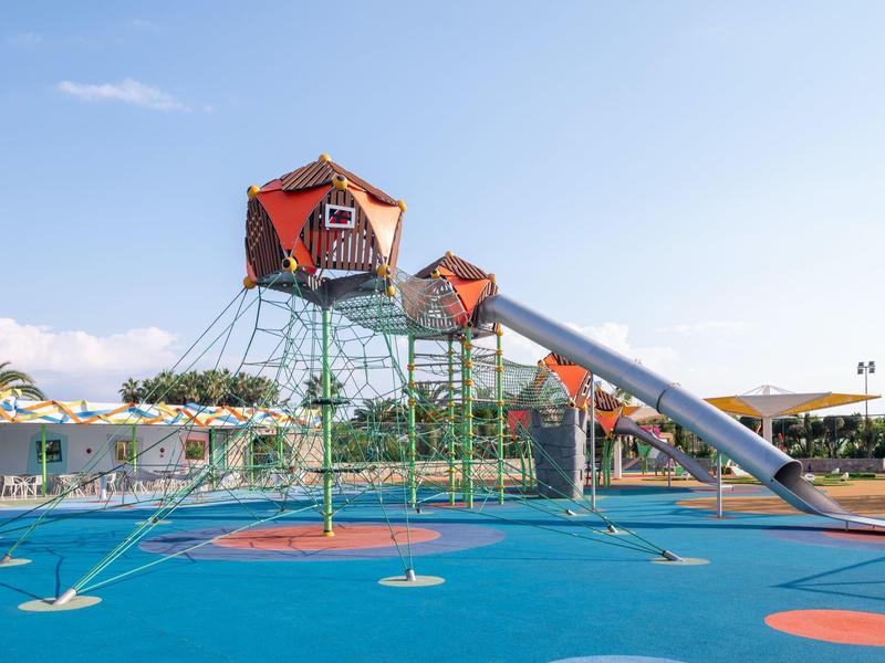 Parque infantil con torres futuristas y un tobogán largo sobre suelo azul en un día soleado.