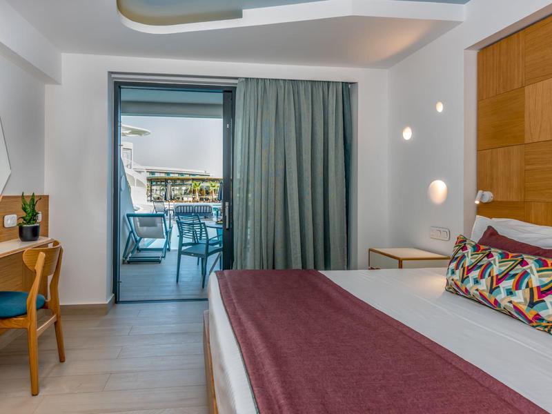 Habitación de hotel luminosa con cama, cojín colorido, escritorio y vista a terraza con sillas.