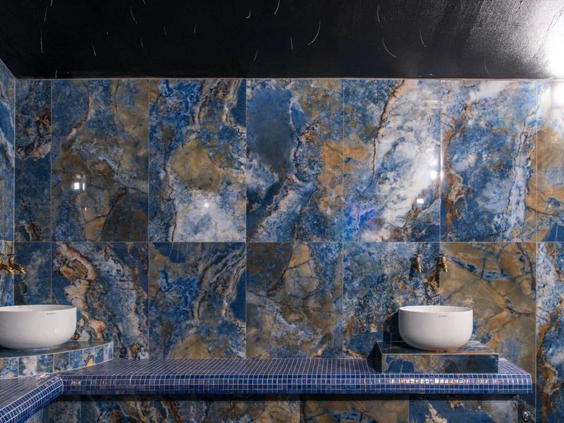 Pared de baño con mármol azul y dorado y dos lavabos blancos sobre mosaicos azules.