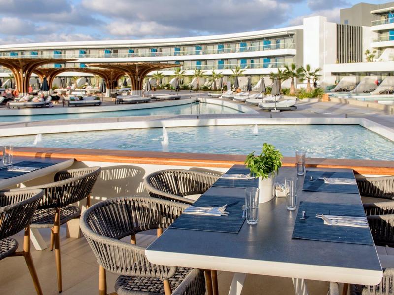 Terraza del hotel con mesas, sillas y piscina frente a un edificio moderno bajo un cielo nublado.