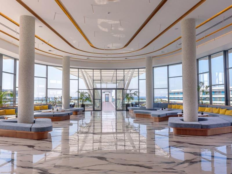 Gran vestíbulo de hotel luminoso con suelo de mármol, columnas y áreas de descanso modernas con vistas al exterior.