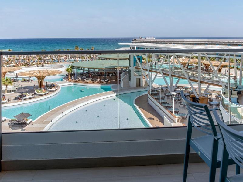 Vista desde el balcón con silla hacia una piscina de hotel y el mar bajo un cielo despejado.