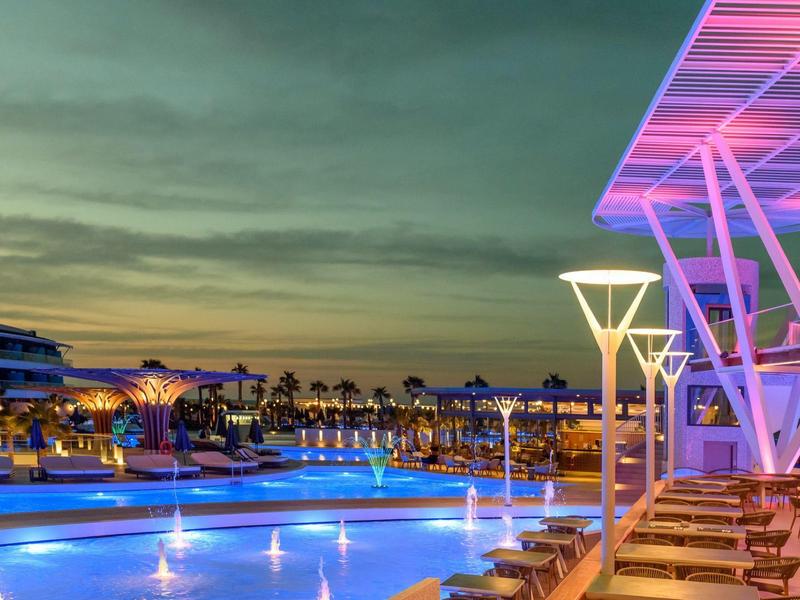 Piscina iluminada con luces azules junto a un área de descanso bajo una estructura moderna al atardecer.