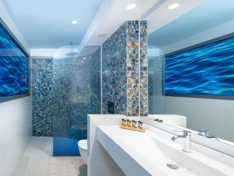 Baño moderno con azulejos de mosaico, espejo grande y decoración azul en la habitación del hotel.