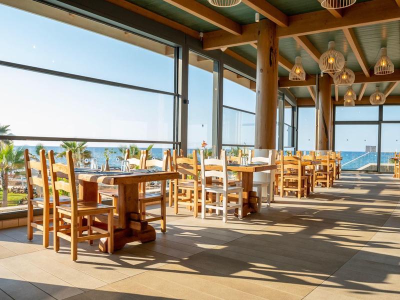 Restaurante luminoso y abierto con muebles de madera y vista al mar y palmeras.