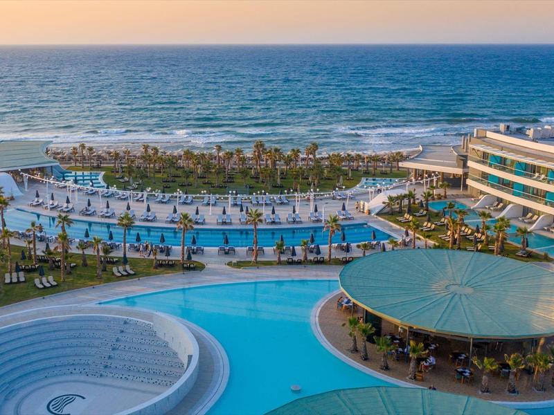 Complejo hotelero de lujo frente al mar con varias piscinas, palmeras y edificios modernos al atardecer.
