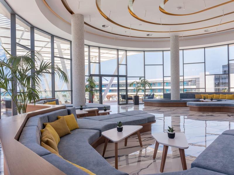 Sala luminosa con grandes ventanales, sofás circulares y vistas a la terraza del hotel y al mar.