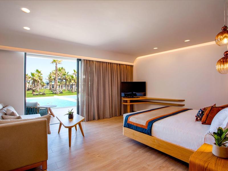 Habitación de hotel moderna con cama grande, zona de estar y vista a la piscina y palmeras.