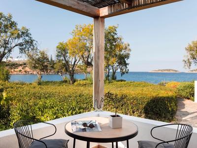 Terrasse avec table et chaises, vue sur des plantes vertes et la mer bleue en arrière-plan.