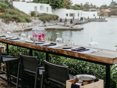 Table en bois avec chaises face à la mer et des maisons blanches en arrière-plan.
