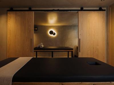 Une table de massage sombre devant un mur avec des panneaux de bois chaleureux et un éclairage minimaliste.
