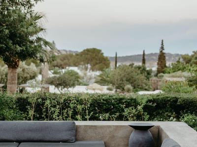 Espace lounge avec canapés gris et tabourets devant un jardin verdoyant sous un ciel dégagé.