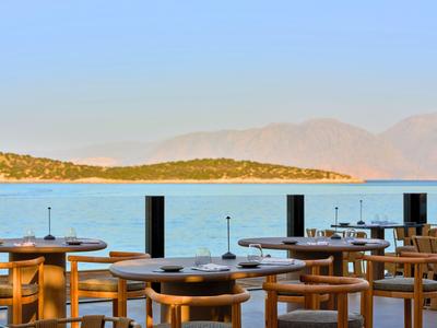 Espace de restaurant ouvert avec chaises en bois et tables donnant sur une mer calme et des montagnes.