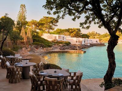 Terrasse avec tables et chaises en bois au bord de l'eau devant des bâtiments blancs dans une baie calme.