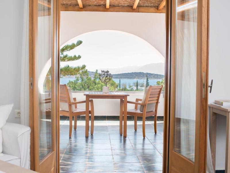 Vue à travers des portes en bois ouvertes sur une terrasse avec table, chaises et vue sur la mer par ciel clair.