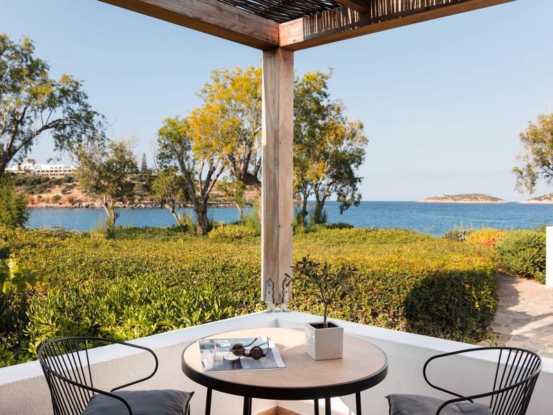 Terrasse avec table et chaises, vue sur des plantes vertes et la mer bleue en arrière-plan.
