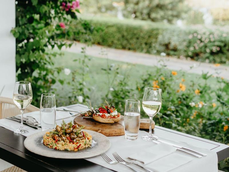 Table en plein air avec vin et plats méditerranéens, entourée de plantes vertes.