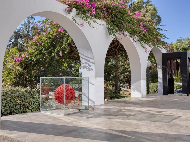 Arches blanches avec fleurs roses et œuvre d'art rouge dans un jardin ensoleillé.