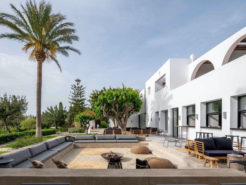 Bâtiment d'hôtel blanc avec terrasse, piscine et paysage tropical de palmiers sous un ciel bleu.