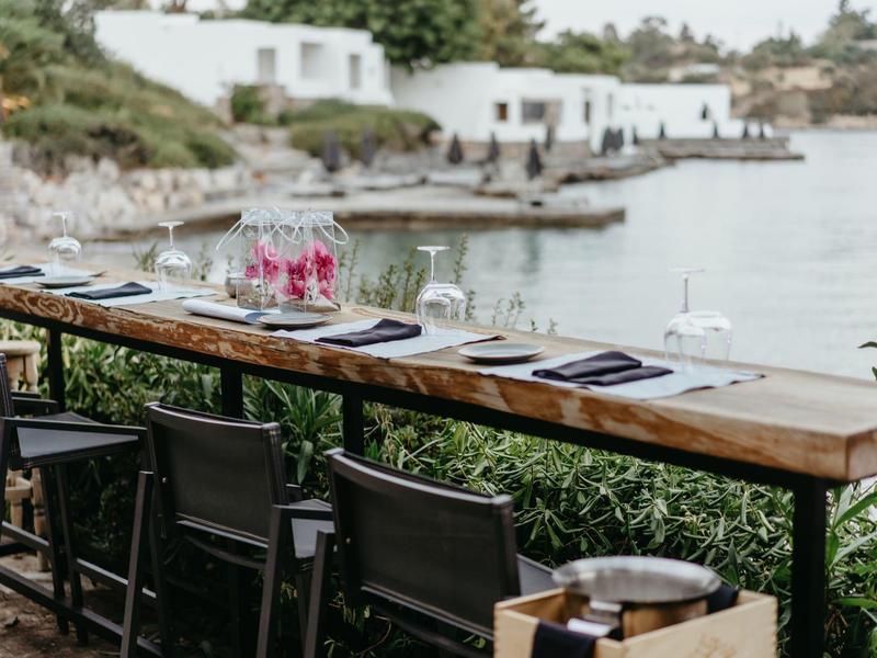 Table en bois avec chaises face à la mer et des maisons blanches en arrière-plan.