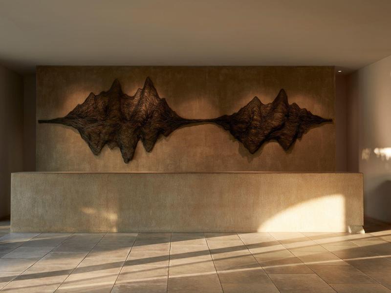 Sculpture murale en métal foncé dans un hall aux tons neutres éclairé par la lumière naturelle.