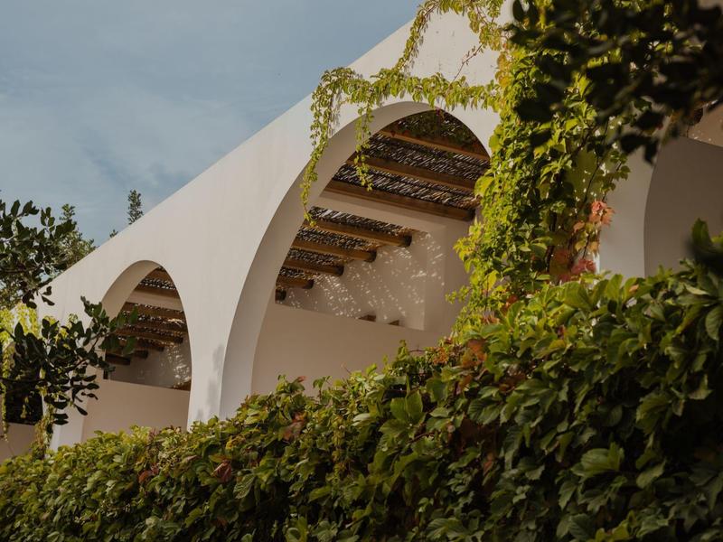 Bâtiments blancs avec des arches et beaucoup de verdure au premier plan sous un ciel nuageux.