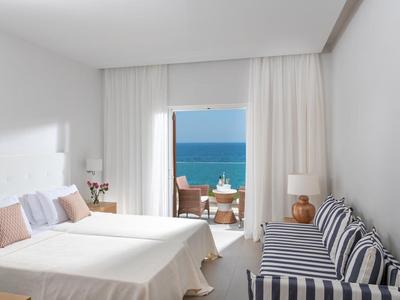 Schlichtes, helles Zimmer mit Doppelbett, gestreiftem Sofa und Balkon mit Meerblick.