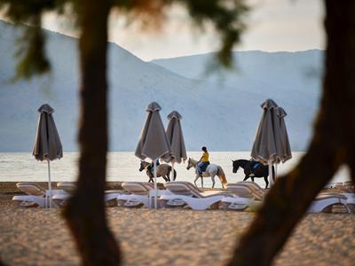 Drei Kinder reiten am Strand zwischen geschlossenen Sonnenschirmen, im Hintergrund Berge und Meer sichtbar.