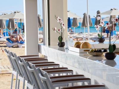 Moderner Strandbar-Tresen in Weiß mit Hockern, Orchideen-Deko und Strand mit Sonnenschirmen im Hintergrund.
