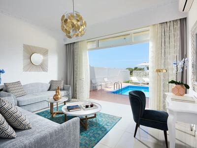Heller Wohnraum mit Sofa, Teppich, Sesseln und bodentiefen Fenstern mit Blick auf Pool und Terrasse.