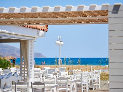 Freiluftrestaurant mit weißen Stühlen und Tischen, blauem Meer und klarem Himmel im Hintergrund.