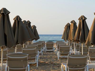 Reihe geschlossener Sonnenschirme und Stühle am Strand mit ruhigem Meer und klarem Himmel.