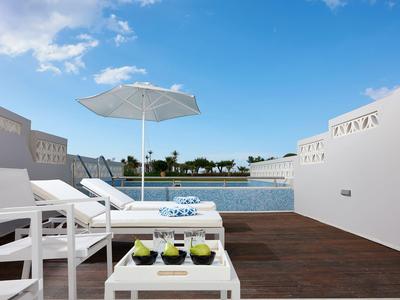 Terrasse mit weißen Liegestühlen, Sonnenschirm, Tisch mit Getränken und Blick auf Wasser und Häuser.