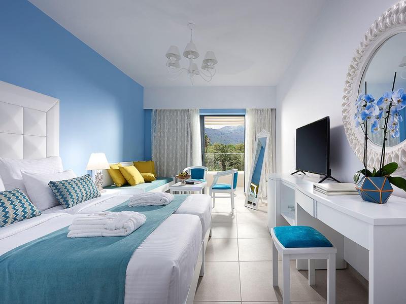 Bright hotel room with two beds, blue and white decor, and sea view through the window.