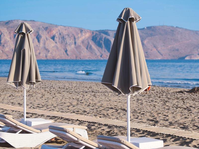Closed gray beach umbrellas and lounge chairs on sandy beach with sea and mountains in background.