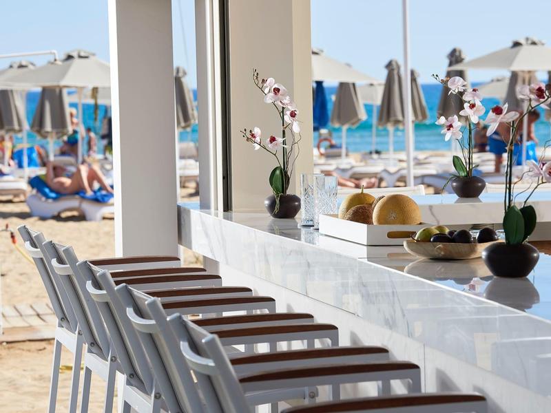 Bar area with chairs overlooking a sandy beach with umbrellas and lounge chairs.