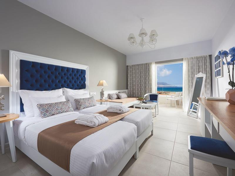 Modern hotel room with large beds, balcony, and sea view in daylight.