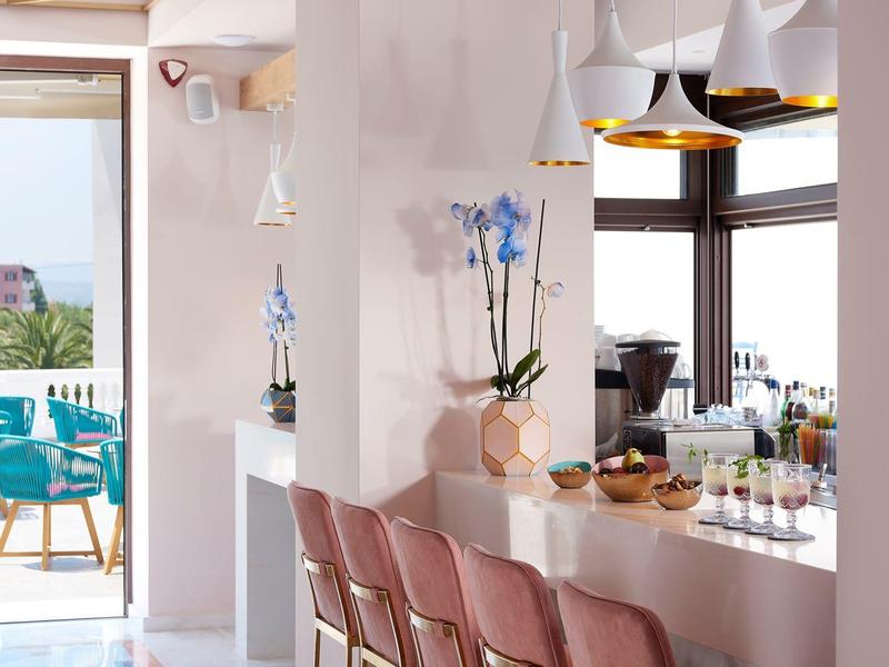 Modern hotel bar area with pink chairs, blue flowers, and view of terrace and sea.