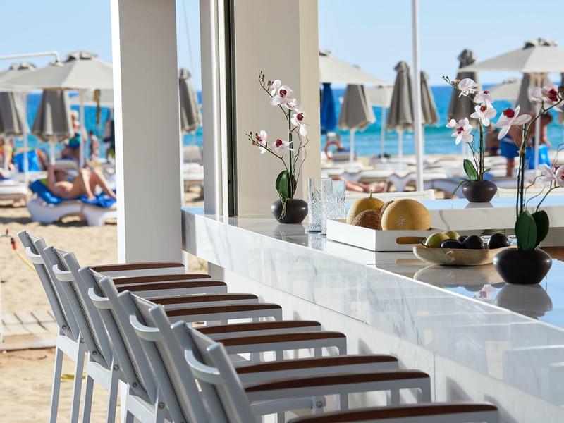 Modern bar with white chairs overlooking beach umbrellas and sun loungers on the sand.