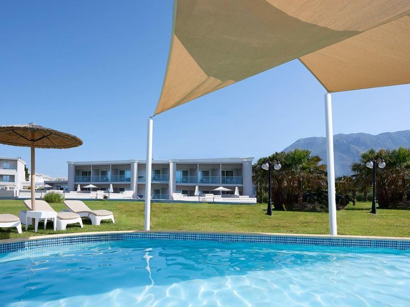 View of a pool with lounge chairs and modern hotel buildings under sunshades.