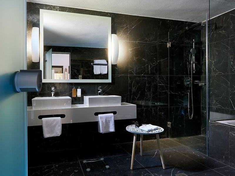 Modern hotel bathroom with double sinks, large mirror, and dark tile walls.