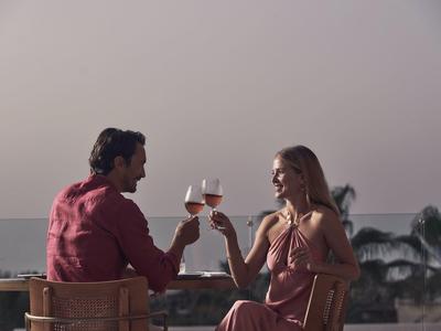 Paar genießt bei Sonnenuntergang Wein auf Terrasse mit Meerblick und Palmen im Hintergrund.